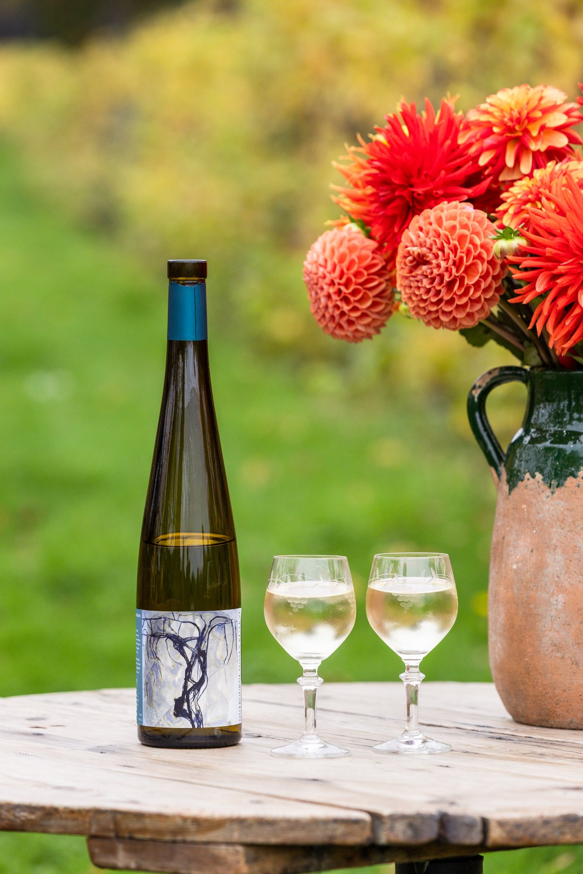 A bottle of Nutbourne Sussex Reserve beside two filled wine glasses on a wooden table, with dahlias and the vineyard behind