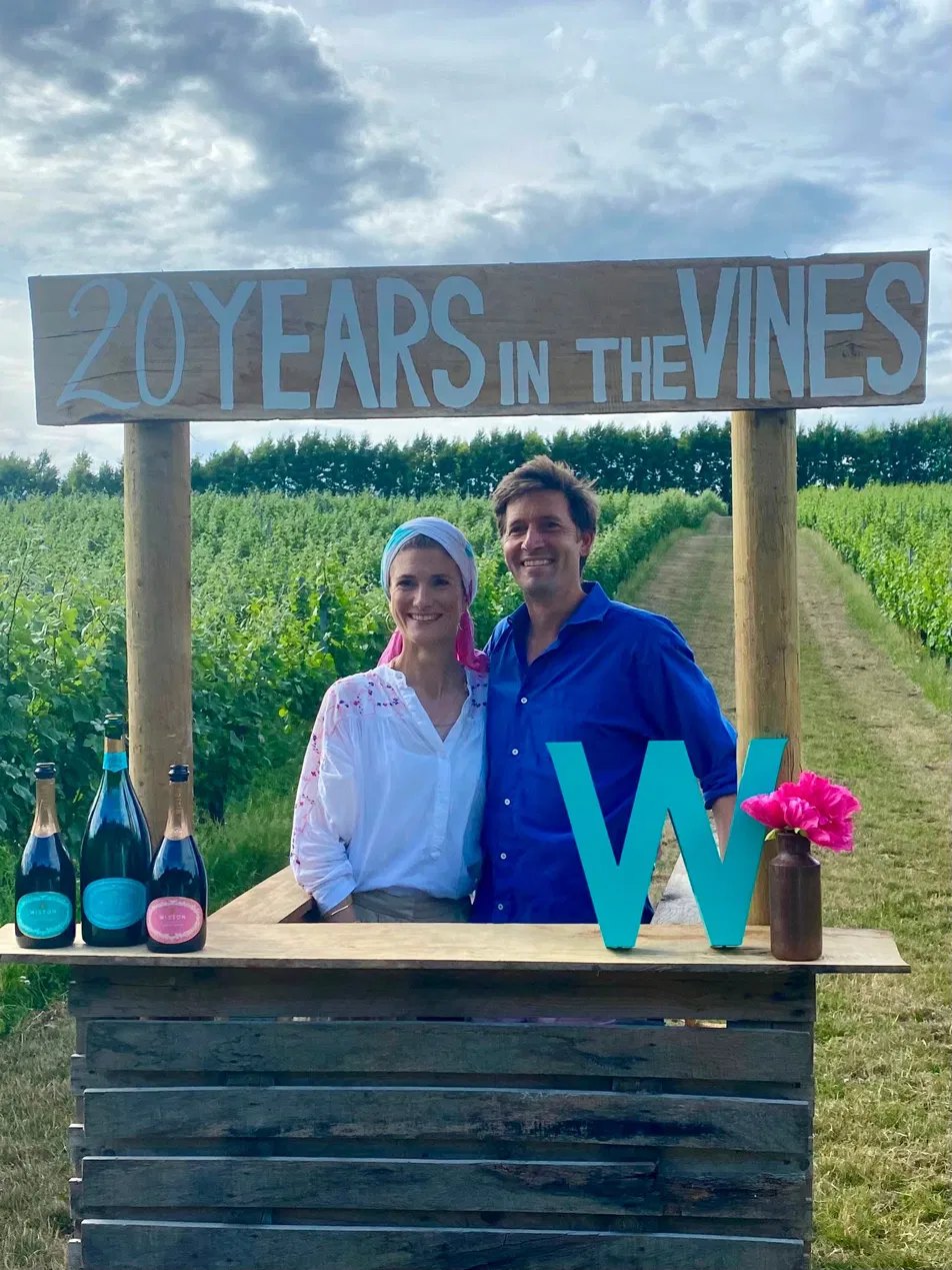 A hand-painted '20 Years in the Vines' sign above a wooden bar set among the vineyard rows at Wiston Estate, with bottles of Wiston sparkling wine on the counter